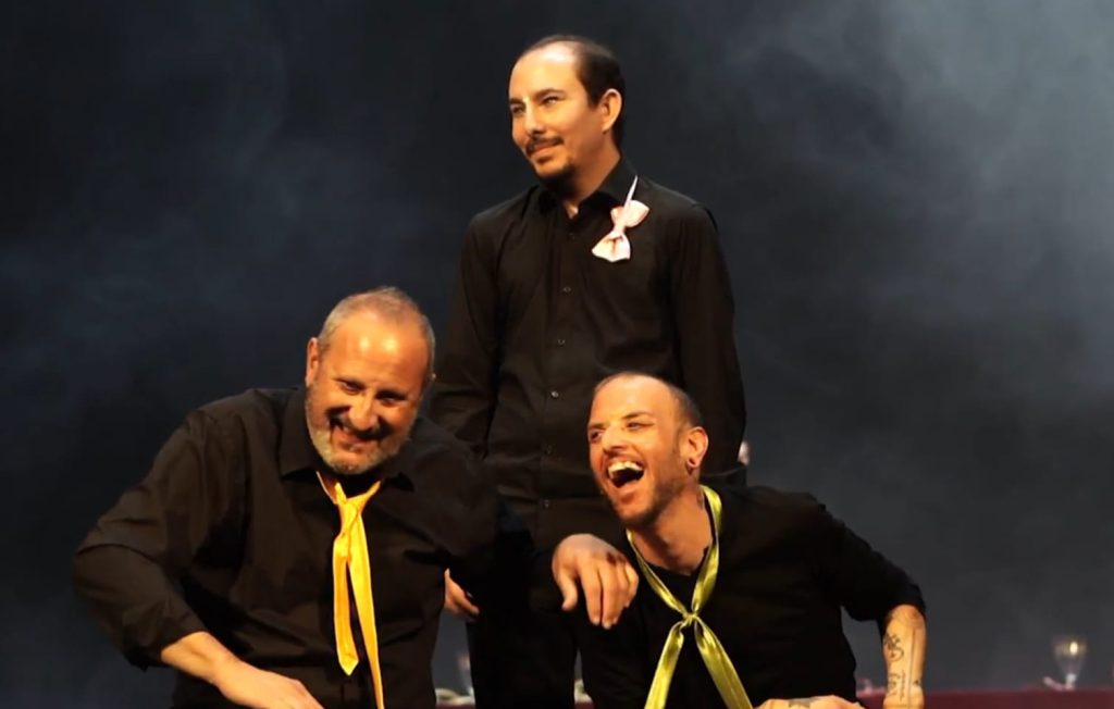 La imagen muestra a tres integrantes de In-Visible Teatro durante la representación de su obra 3 locos, 2 enamorados, 1 boda, 1 funeral y Ramsés..., sobre un escenario con fondo oscuro e iluminación cálida que destaca las figuras. Los intérpretes visten de negro y llevan elementos llamativos, como corbatas de colores y un detalle decorativo en el pecho, que aportan contraste visual. La proximidad entre los cuerpos y la disposición de las posturas crean una composición dinámica que refleja el tono escénico de la representación.