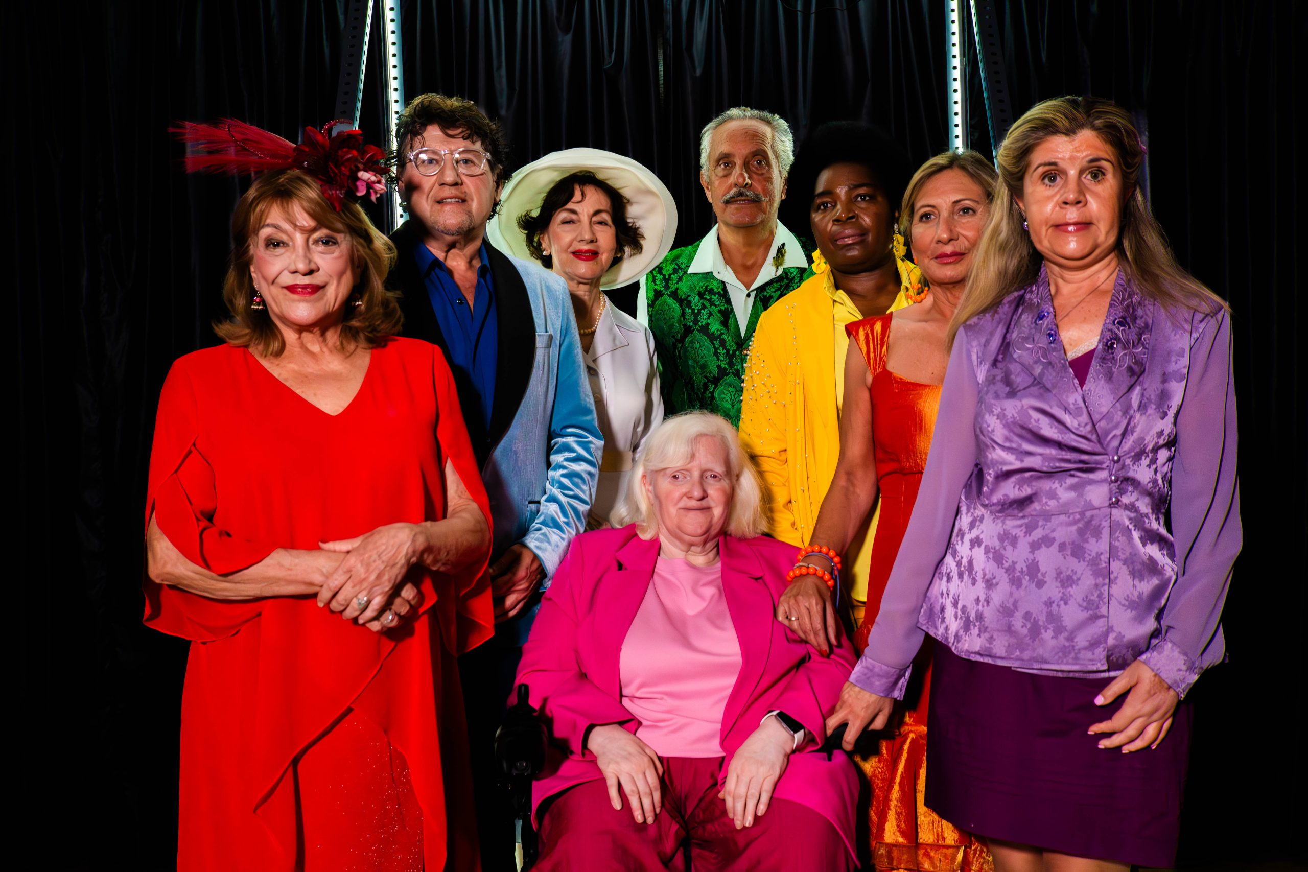 La imagen muestra al elenco de la agrupación teatral El Malecón posando en un escenario con fondo oscuro, luciendo vestuario variado y colorido que destaca visualmente. En el centro, una persona aparece sentada, rodeada por el resto del grupo de pie, formando una composición colectiva equilibrada. El conjunto transmite el carácter coral y escénico de la comedia El Malecón 9ºB, ambientada en situaciones humorísticas dentro de un edificio.