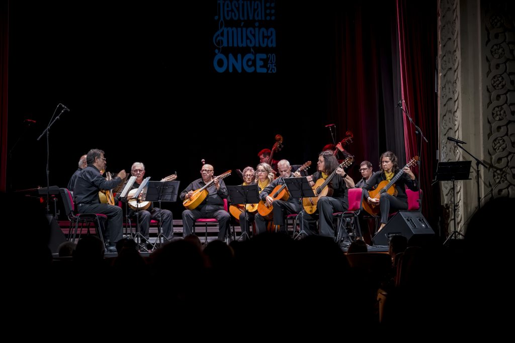 La imagen muestra a la Orquesta Ciudad de los Califas actuando en un escenario durante el Festival de Música de la ONCE 2025, con iluminación centrada sobre los músicos y fondo oscuro. En el centro, varias personas interpretan instrumentos de cuerda como guitarras, laúdes y bandurrias, mientras una persona dirige desde el lateral izquierdo con ayuda de una partitura. El conjunto está rodeado de micrófonos y atriles, con el público visible en la parte inferior, creando un ambiente de concierto formal y en directo.