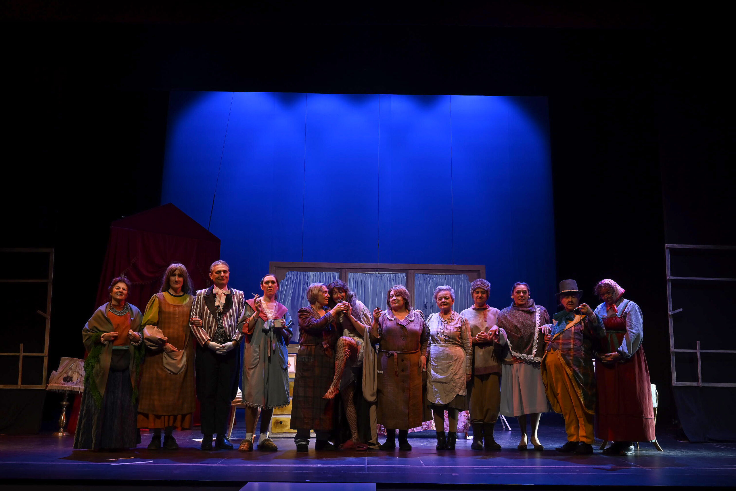 El elenco de la agrupación Jacaranda aparece alineado sobre el escenario tras la función, ataviado con vestuario teatral de época variado y detallado.
El fondo muestra un decorado escénico con una estructura doméstica y una intensa iluminación azul que envuelve la escena.
La imagen captura el momento final colectivo tras el estreno de Tormenta de Otoño, con todos los intérpretes reunidos en el cierre.