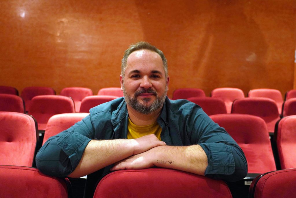 Jamp Paló, director artístico de El Malecón, aparece apoyado en una butaca dentro de un teatro con filas de asientos rojos, en un entorno claramente vinculado a la creación escénica.
La escena refleja una estética contemporánea y cercana, acorde con su impulso renovador basado en el humor, el ritmo y el simbolismo.